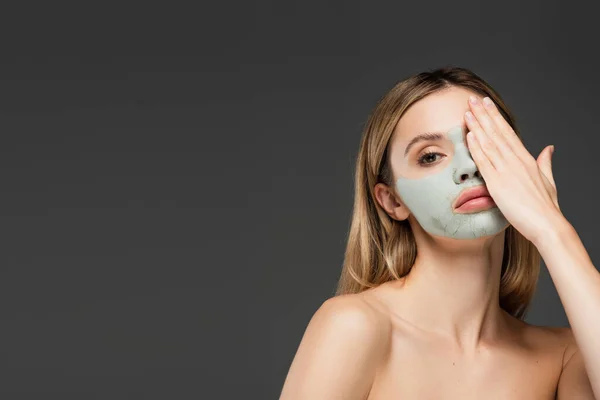 Jeune femme aux épaules nues et masque d'argile visage obscurcissant à la main isolé sur gris — Photo de stock