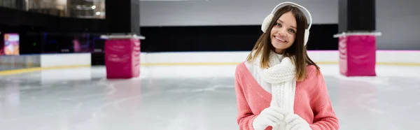 Cheerful young woman in ear muffs and scarf on ice rink, banner — Stock Photo