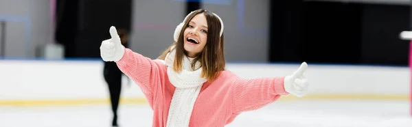 Mujer joven feliz en orejeras y bufanda que muestra los pulgares hacia arriba en la pista de hielo, bandera - foto de stock