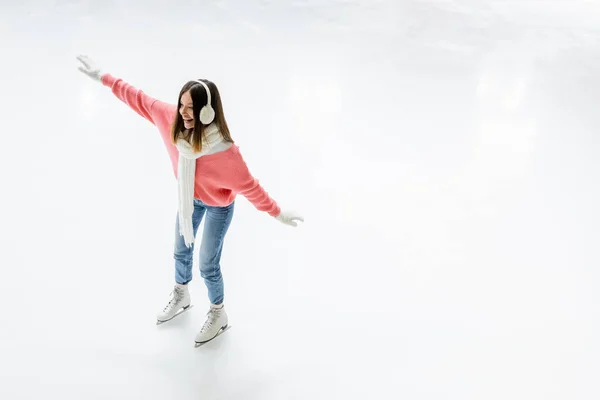 Vista de ángulo alto de la joven feliz en orejeras y bufanda patinaje en pista de hielo - foto de stock