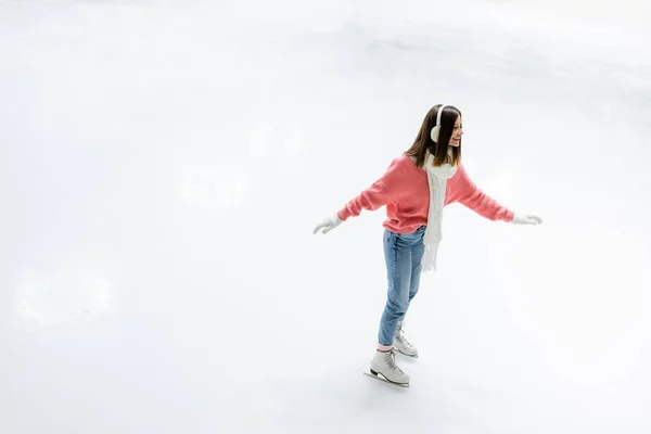 Vue grand angle de la jeune femme gaie dans des cache-oreilles et foulard patiner sur la patinoire — Photo de stock