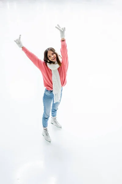 Vue grand angle de la jeune femme joyeuse dans des cache-oreilles et une écharpe montrant signe de paix tout en patinant sur la patinoire — Photo de stock