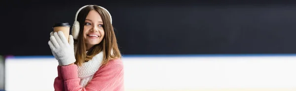 Cheerful young woman in ear muffs and scarf holding paper cup on ice rink, banner — Stock Photo