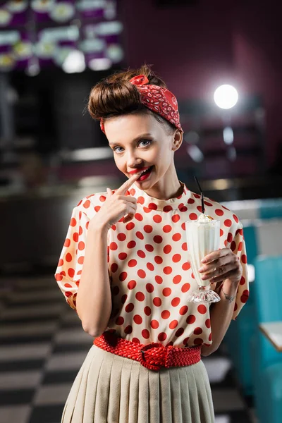 Feliz pin up mulher segurando milkshake em vidro e lambendo o dedo no café — Fotografia de Stock