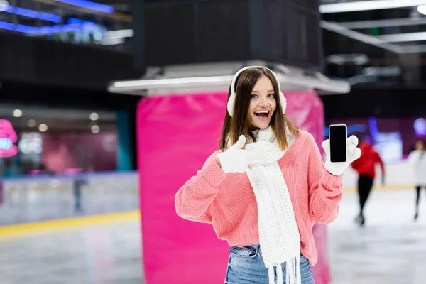 Изумленная женщина в белых наушниках держит смартфон с пустым экраном и показывает большой палец на катке — Stock Photo