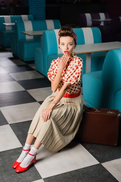 Surprise pin up femme en robe assis près de valise rétro dans le café — Photo de stock