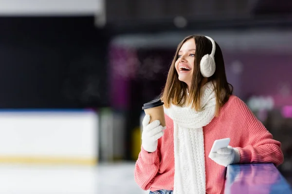 Donna eccitata in paraorecchie tenendo tazza di carta e smartphone sulla pista di pattinaggio su ghiaccio — Foto stock