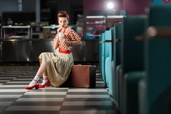Молодая женщина, пьющая молочный коктейль и сидящая на винтажном чемодане в кафе — стоковое фото