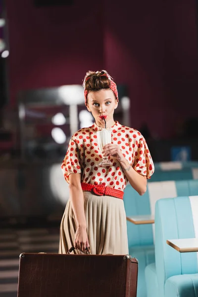 Pin up woman holding suitcase and drinking tasty milkshake in cafe — Stock Photo