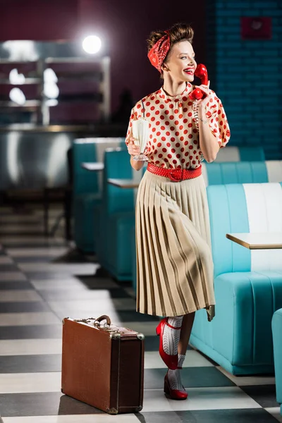 Happy pin up woman talking on telephone and holding tasty milkshake near retro suitcase in cafe — Stock Photo