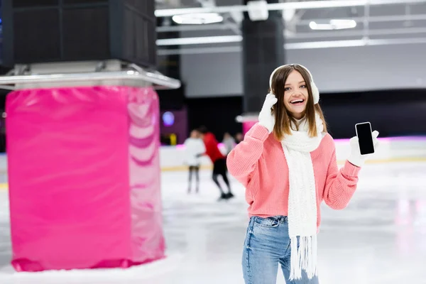 Femme étonnée dans des cache-oreilles blancs tenant smartphone avec écran blanc sur la patinoire — Photo de stock