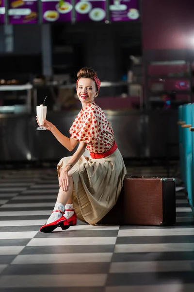 Happy pin up woman holding milkshake while sitting on retro suitcase in cafe — Stock Photo