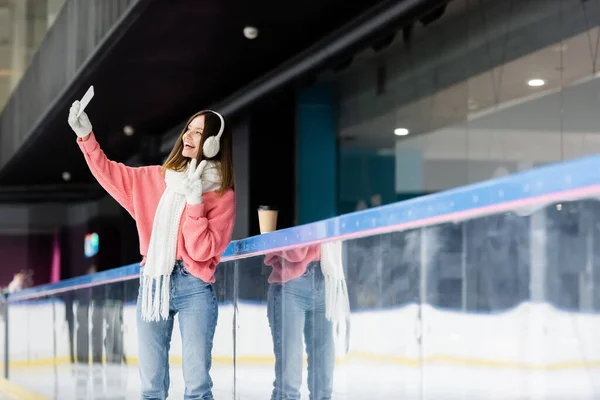 Mujer joven positiva tomando selfie mientras que muestra la muestra de la paz cerca de taza de papel en pista de hielo - foto de stock