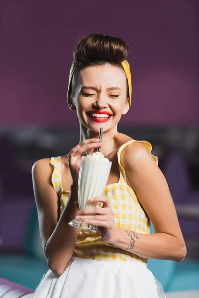 Alegre pin up mujer con los ojos cerrados sosteniendo vaso de sabroso batido en la cafetería - foto de stock