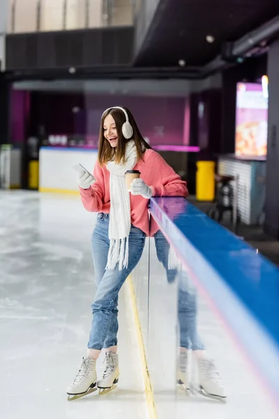 Full length of young woman with paper cup and smartphone standing on ice rink — Stock Photo