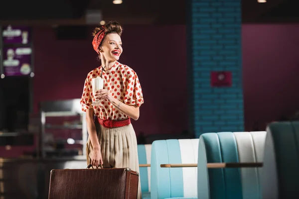 Positive pin up woman holding suitcase and cocktail in cafe — Stock Photo