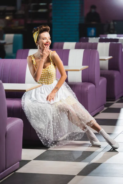 Longitud completa de la mujer pin up alegre sentado en el asiento púrpura en la cafetería - foto de stock