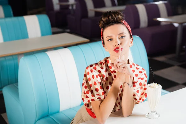 Jovem pin up mulher mostrando sinal de silêncio perto de milkshake na mesa — Fotografia de Stock