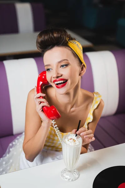 Положительный пин-ап женщина говорит по телефону и держит питьевой соломы возле молочного коктейля — стоковое фото