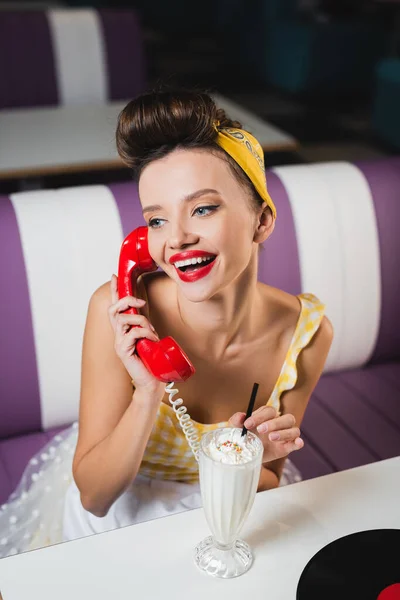 Positivo pin up mujer hablando por teléfono y la celebración de beber paja cerca de batido - foto de stock