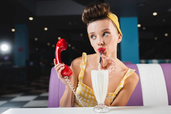 Joven pin up mujer bebiendo batido y sosteniendo el auricular rojo - foto de stock