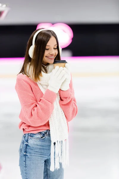 Happy woman in white gloves and ear muffs holding paper cup on ice rink — Stock Photo