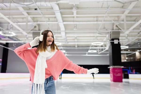 Mulher animada em abafadores de ouvido e suéter rosa patinação na pista de gelo — Fotografia de Stock