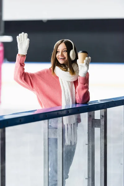 Mulher animada em muffs ouvido e suéter rosa segurando copo de papel e mão acenando na pista de gelo — Fotografia de Stock