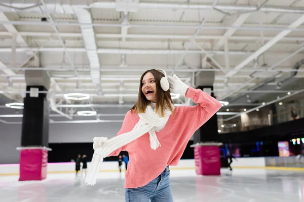 Femme excitée dans des cache-oreilles et pull rose sur patinoire gelée — Photo de stock