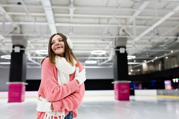 Pleased woman in ear muffs and pink sweater embracing herself on ice rink — Stock Photo