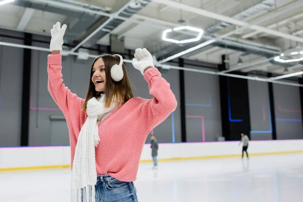 Donna eccitata in manicotti auricolari e maglione rosa con le mani sollevate sulla pista di ghiaccio — Foto stock