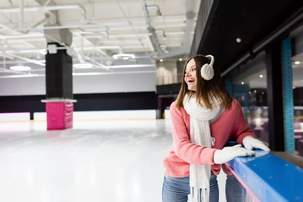 Femme excitée dans des cache-oreilles, écharpe et pull rose sur la patinoire — Photo de stock