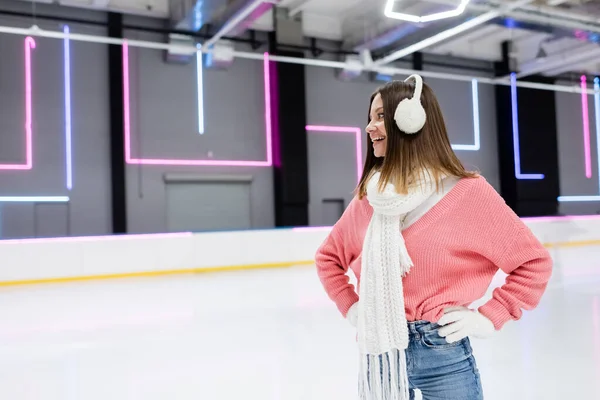 Mujer feliz en orejeras, bufanda y suéter rosa de pie con las manos en las caderas en la pista de hielo - foto de stock