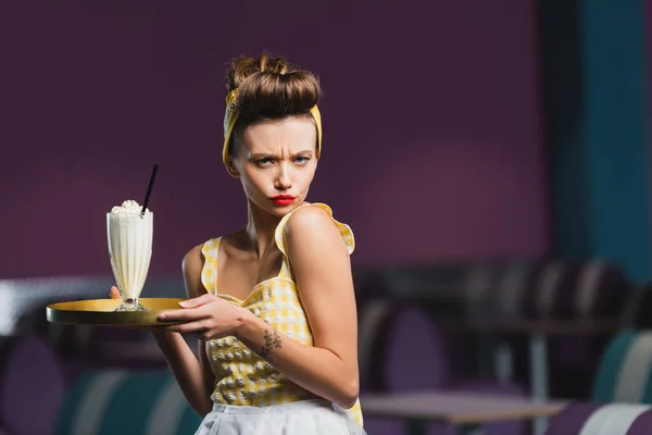 Descontento pin up camarera con tatuaje celebración bandeja y batido en la cafetería - foto de stock