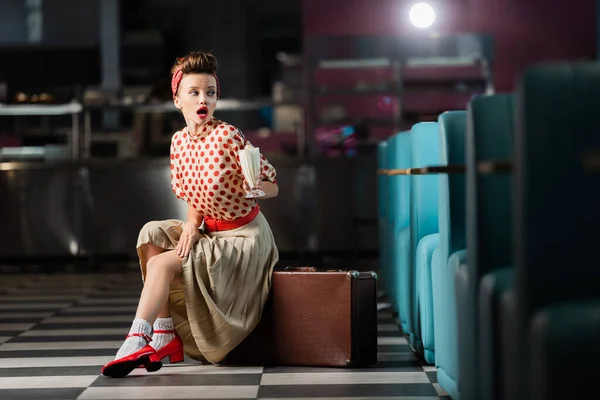 Surprised pin up woman holding milkshake cocktail and sitting on suitcase in cafe — Stock Photo