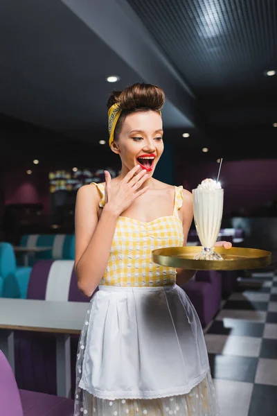 Asombrado pin up camarera mirando bandeja con batido en la cafetería - foto de stock