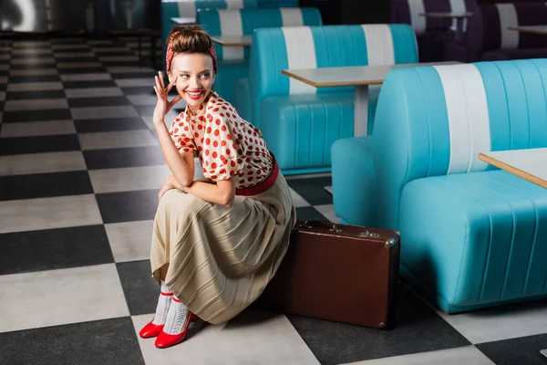 Happy pin up woman sitting on vintage suitcase in cafe — Stock Photo