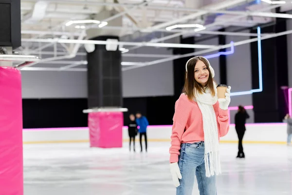 Happy woman in ear muffs and pink sweater holding paper cup on ice rink — Stock Photo