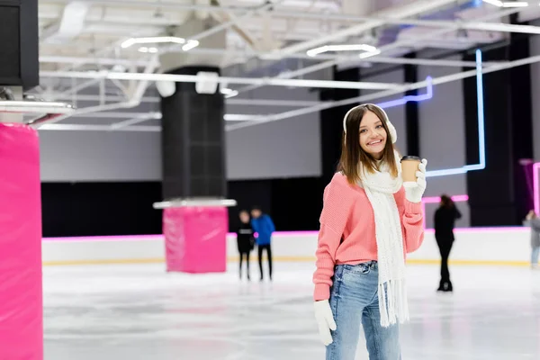 Femme heureuse dans des cache-oreilles et pull rose tenant tasse en papier sur la patinoire — Photo de stock