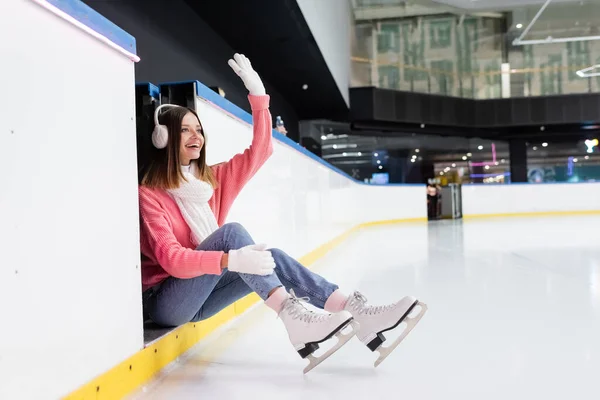 Mujer feliz en orejeras y suéter rosa saludando mano en pista de hielo - foto de stock