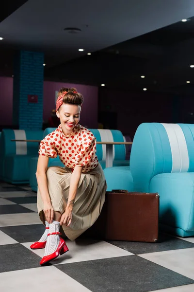 Happy pin up woman sitting on vintage suitcase and adjusting sock in cafe — Stock Photo