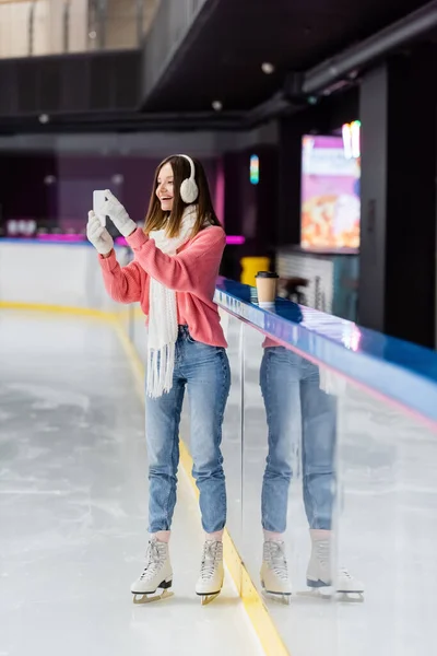 Full length of brunette woman in gloves and ear muffs taking photo on ice rink — Stock Photo