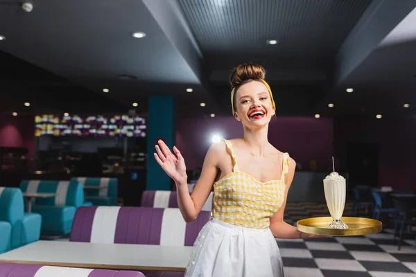 Emocionado joven pin up camarera sosteniendo bandeja con batido y gesto en la cafetería - foto de stock