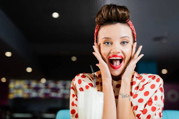 Aufgeregte Pin-up-Frau blickt in Kamera neben verschwommenem Milchshake-Glas — Stockfoto