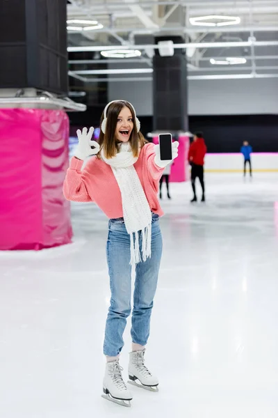 Excited woman in gloves and ear muffs showing ok sign while holding smartphone with blank screen on ice rink — Stock Photo