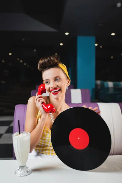 Happy pin up woman with red lips holding vinyl disc and talking on telephone near milkshake in cafe — Stock Photo