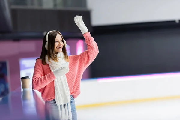 Happy woman in ear muffs holding smartphone and waving hand on ice rink — Stock Photo