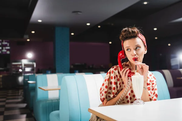 Young pin up woman drinking milkshake and talking on retro telephone — Stock Photo