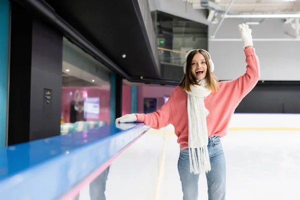 Jeune femme excitée en pull rose, écharpe et cache-oreilles agitant la main sur la patinoire — Photo de stock