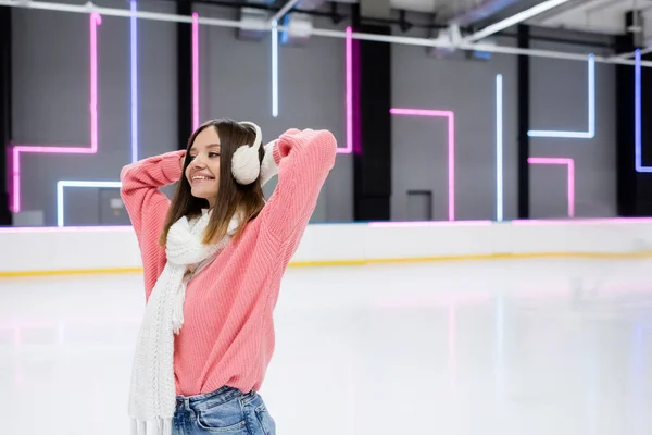 Sorrindo jovem mulher em suéter rosa, cachecol e abafadores de ouvido na pista de gelo — Fotografia de Stock